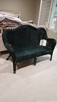 Black rattan wicker loveseat with visible wear, scratches, peeling paint and some breaks shown from front angle in interior setting.