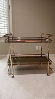 Front view of rolling bar cart showing metal frame, top and bottom glass shelves, and wheels.