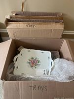 Open cardboard box containing a white ceramic serving tray with floral designs on handles and center, surrounded by bubble wrap and additional boxes labeled 'TRAYS'