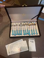Photo showing full set of 6 dessert spoons and 6 dessert forks in original brown case with satin lined interior. Cloisonne handles are turquoise blue with white floral pattern. Certificates of authenticity visible outside the case.