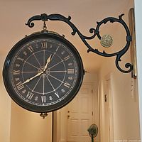 Front angle view of mounted double-sided wall clock showing black face with Roman numerals and ornate scrollwork metal bracket with decorative pendulum.