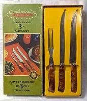 Photo of the Galaxie Stainless Hollow Ground 3-piece carving set in original packaging with yellow insert showing carving knife, boning knife, and carving fork with tortoiseshell-style handles.