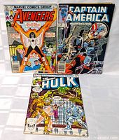 Three vintage Marvel comic books standing upright in protective plastic sleeves on white fabric. Avengers #227 on the left, Captain America #247 in center back, and The Incredible Hulk #273 at front.
