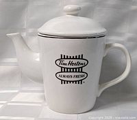 Front view of white ceramic coffee/tea pot with handle and lid showing Tim Hortons logo.