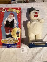 Hip Swinging Santa doll displayed in original festive box and white snowman plush figure standing beside it on blue base.