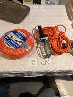 View of the new coiled Mastercraft 30m extension cord and the coiled orange trouble light with protective metal cage and hook on a white surface.