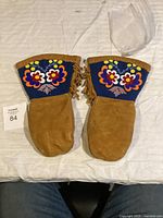 Pair of mittens laid out on a white cloth, showing front sides with brown suede leather and blue cuffs with colorful floral beadwork and embroidery.