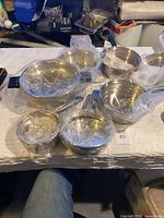 Top-down view of various stainless steel cookware pieces wrapped in plastic on a white cloth surface.