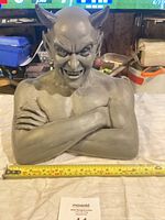 Front view of gray devil bust statue with horns, crossed arms, and intense facial expression, measuring about 15 inches wide with tape measure shown.