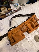 Full view of the brown leather tool belt laid flat, showing multiple pockets and black adjustable belt strap.