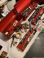 Overview of three red metal toolboxes open, revealing various hand tools and hardware items inside and around them on a white surface.