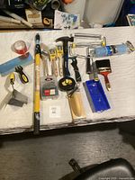 Overview of all tools including paintbrushes, roller handles, hammer, tape measure, putty knife, adhesive tape, drill bits, and paint tray on white surface.
