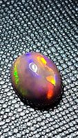 Close-up image of oval cabochon natural black opal on a textured black background showing bright flashes of rainbow colors.