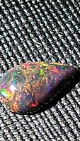 Close-up photo of the pear-shaped black opal gemstone showing multicolor fire and flash on a textured dark fabric background.