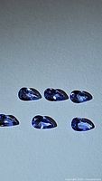 Six small pear-shaped blue purple Tanzanite gemstones arranged on a plain background, showing their calibrated uniform size and cut.