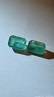 Close-up photo of two octagon cut emerald gemstones showing green color and natural inclusions, lying on plain white surface.