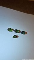 Four natural green tourmaline gemstones lying on a white surface, showing their size, color, and shape (oval and pear).