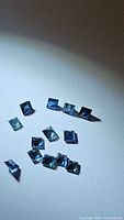 Photo showing multiple princess cut blue to teal green natural sapphires scattered on a white background under light, showcasing their color and cut.