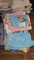 Stack of various vintage linens including aprons, table coverings of different colors and patterns such as blue gingham apron and floral fabrics.