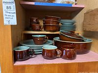Photo showing shelves with stacked stoneware dinnerware items including mugs, plates, bowls, and lidded baking dishes in brown and turquoise glaze.