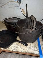 Photo showing two large cast iron pots (one with lid) and a pot containing a cast iron cooking grate, plus a square cast iron skillet on the side.