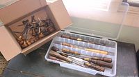 View of wooden handled chisels and screwdrivers in a plastic organizer box and additional tools in a cardboard box on a workbench.