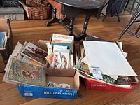 Photo showing two boxes filled with vintage ephemera including newspapers, magazines, cards, and other paper collectibles on a hardwood floor near a wooden table.