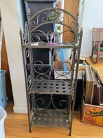 Front view of folding metal shelf showing all three shelves with decorative scrollwork and floral motif on top.