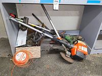 Overall lot showing Husqvarna orange and grey trimmer, green John Deere trimmer behind it, orange trimmer line coil in box, and loose parts on floor beneath shelf.
