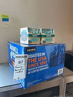Box of Keurig Maxwell House coffee pods and two packs of Renuzit AromaSense candles stacked on a surface against a wall, showing packaging and labels.