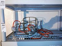 Photo showing two metal cage light fixtures on a grey plastic shelving unit against a white wall. Orange electrical cords are visibly cut and tangled, with mounting attachments attached to each fixture.
