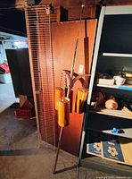 Full view of a tall vintage Mid-Century Modern pole lamp with orange hanging cylindrical lampshades, placed in a basement storage area with surrounding shelving and boards.