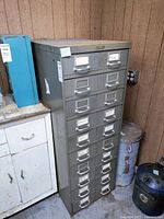 Tall gray vintage metal cabinet with 10 drawers, showing signs of rust and scratches. Handwritten labels on some drawers. Cabinet against wooden wall with other workshop items nearby.