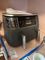 Front view of Ninja Foodi dual basket air fryer on shelf with two pull-out baskets handles in front and digital control panel above them, showing brand name and buttons.