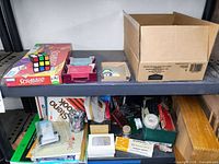 Photo of two shelves containing Scrabble game, Rubik's Cube, several decks of playing cards, assorted paper, notebooks, and some office supplies.