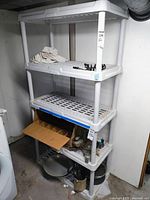 Front view of the white plastic shelving unit in a basement setting showing four lattice shelves, a box, and miscellaneous items stored around it.