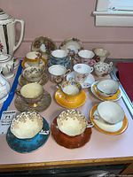 Overview of 14 assorted vintage teacups and saucers arranged on a pink surface with varied floral designs and gold trim, showing different patterns and colors including turquoise, orange, yellow, and white.
