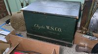 Side view of painted wooden trunk showing brand name 'Clyde W.S. Co.' on dark green paint with black base trim and metal handle.