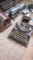Four vintage machines on carpet: Remington typewriter, Protectograph Model H, Paymaster Model Y, Protectograph Model 982911 checkwriters.