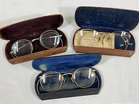 Three pairs of vintage antique eyeglasses in their respective leather covered hard cases arranged side by side on a table, highlighting distinct case colors and interior linings.