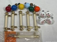 Set of colorful ceramic ball knobs, white ceramic rose floral knobs, and cream drawer handles with brass ends arranged together on a flat surface.