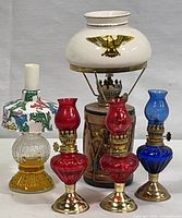 Five oil lamps arranged, showing three smaller colorful glass lamps, the drum-base lamp with eagle motif, and a multicolored glass lamp with plastic shade