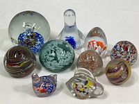 Overview photo of 11 glass paperweights and decorative glass figurines arranged in two rows, showing variety of shapes and colors including a figure with blue and white swirl, multiple round and egg-shaped paperweights with colorful internal designs.
