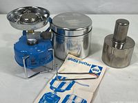 Camping Gaz International blue portable burner with circular top, two interlocking stainless steel pots, detachable handle, instruction sheet, and metal alcohol lamp shown.