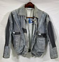 Front view of Guess men's denim jacket on hanger, showing button front, collar, two flap pockets with Guess tags, elastic gathered bottom, grey distressed denim with black leather patches on sleeves and pockets.