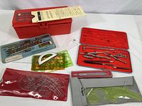 Complete vintage drafting and lettering tool set including metal case, compasses, pens, rulers, protractor, and templates arranged on white background.