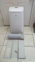Full view of LG portable air conditioner main unit, white housing with LG logo, exhaust hose and window vent pieces on tiled floor