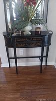 Front view of black Asian style side table with floral arrangement on top, showing one drawer with hand-painted gold and red scenes and bronze hardware.