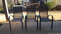 3 black outdoor mesh chairs arranged side by side outside on a patio, framed against an outdoor background including grass and a house.