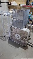 Photo showing the Holmes Brandes box fan, no name box fan, metal stackable shelves, aluminum screen material and metal rolling basket grouped on a garage floor